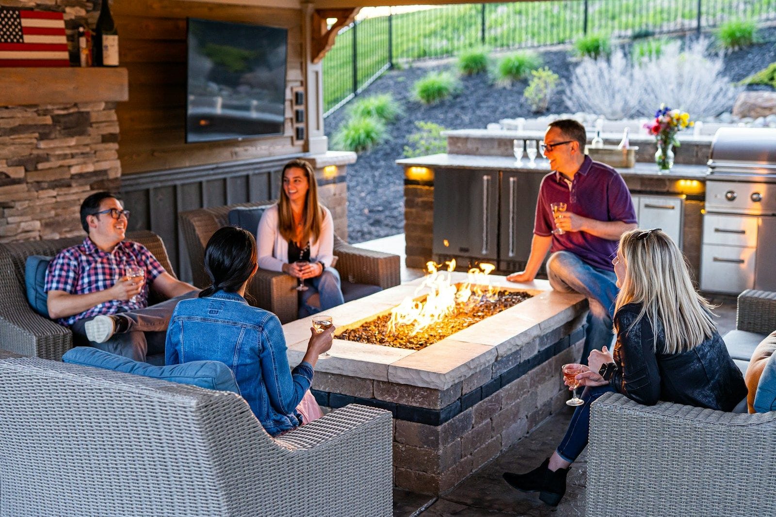 Friends gathered around a gas fire table in a stone-clad outdoor room beside a built-in kitchen.