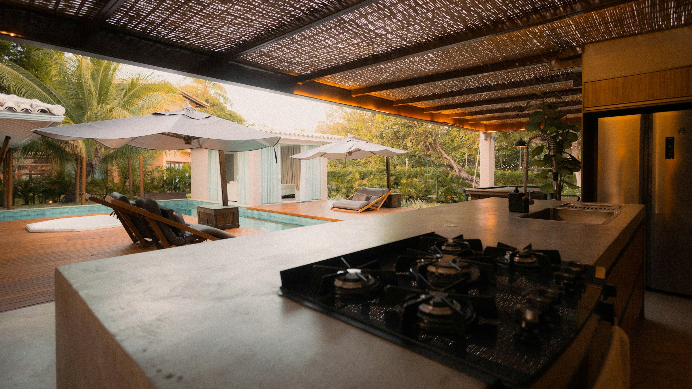 A covered outdoor kitchen with a stone island and cooktop opening onto a timber deck and pool, glowing in warm golden light.