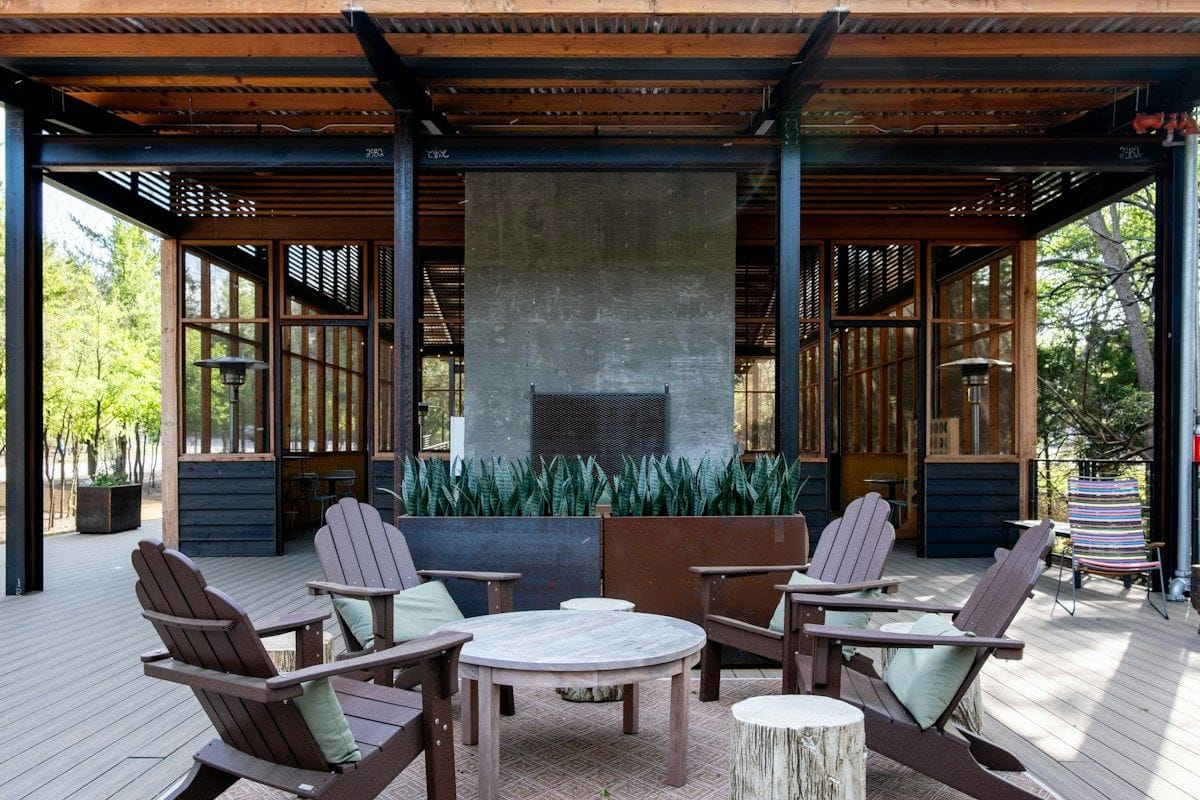 A steel and timber outdoor pavilion with a concrete fireplace, planters and Adirondack chairs around a low table.