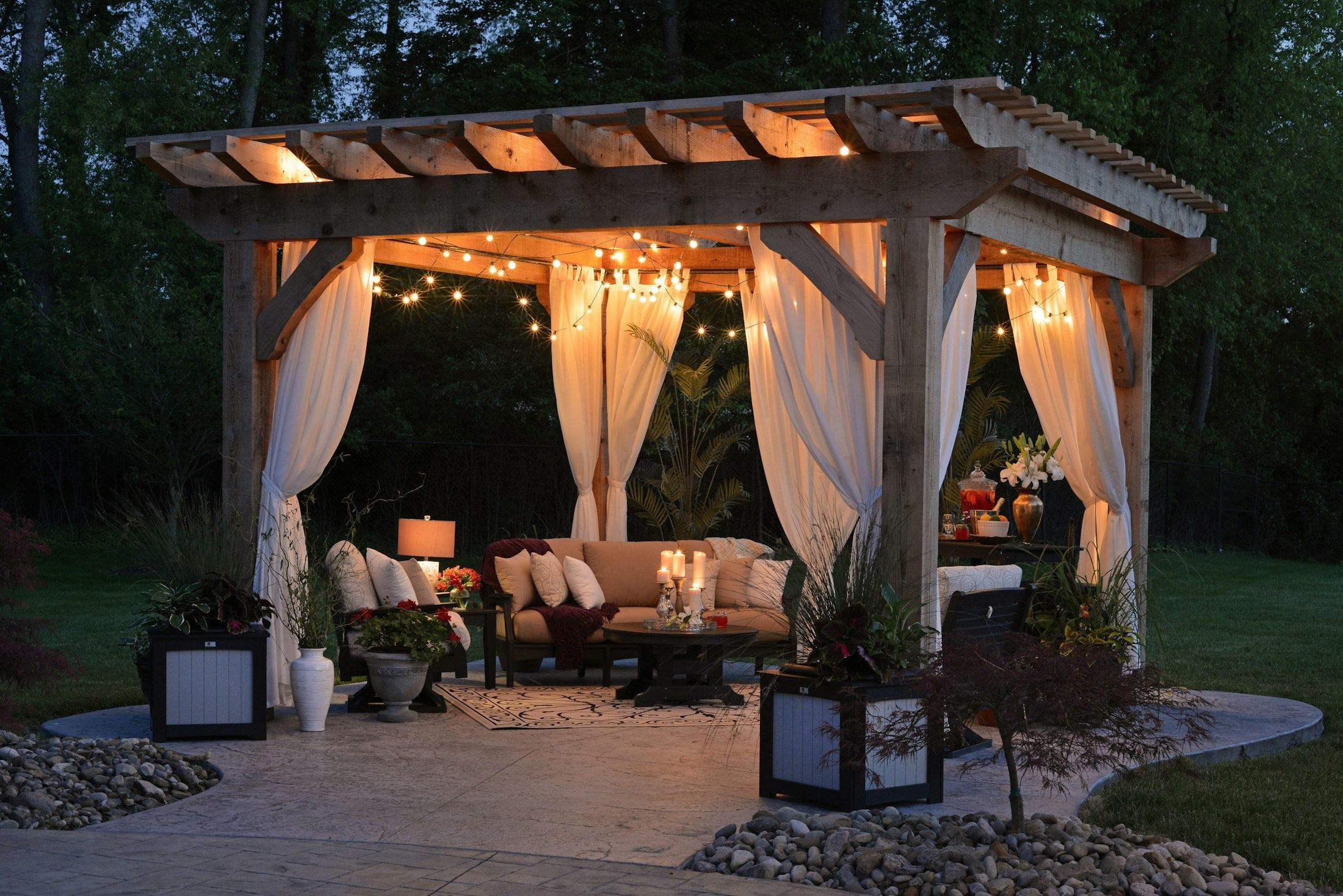 A timber pergola hung with soft curtains and festoon lights over a lounge setting at dusk.